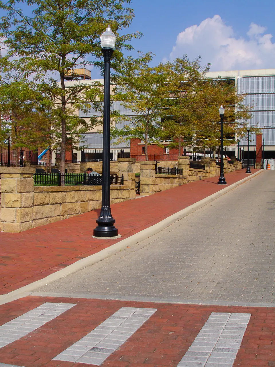 Commercial street edge with brick paving, crosswalk, lamp post, stone walls, lawn, and trees.